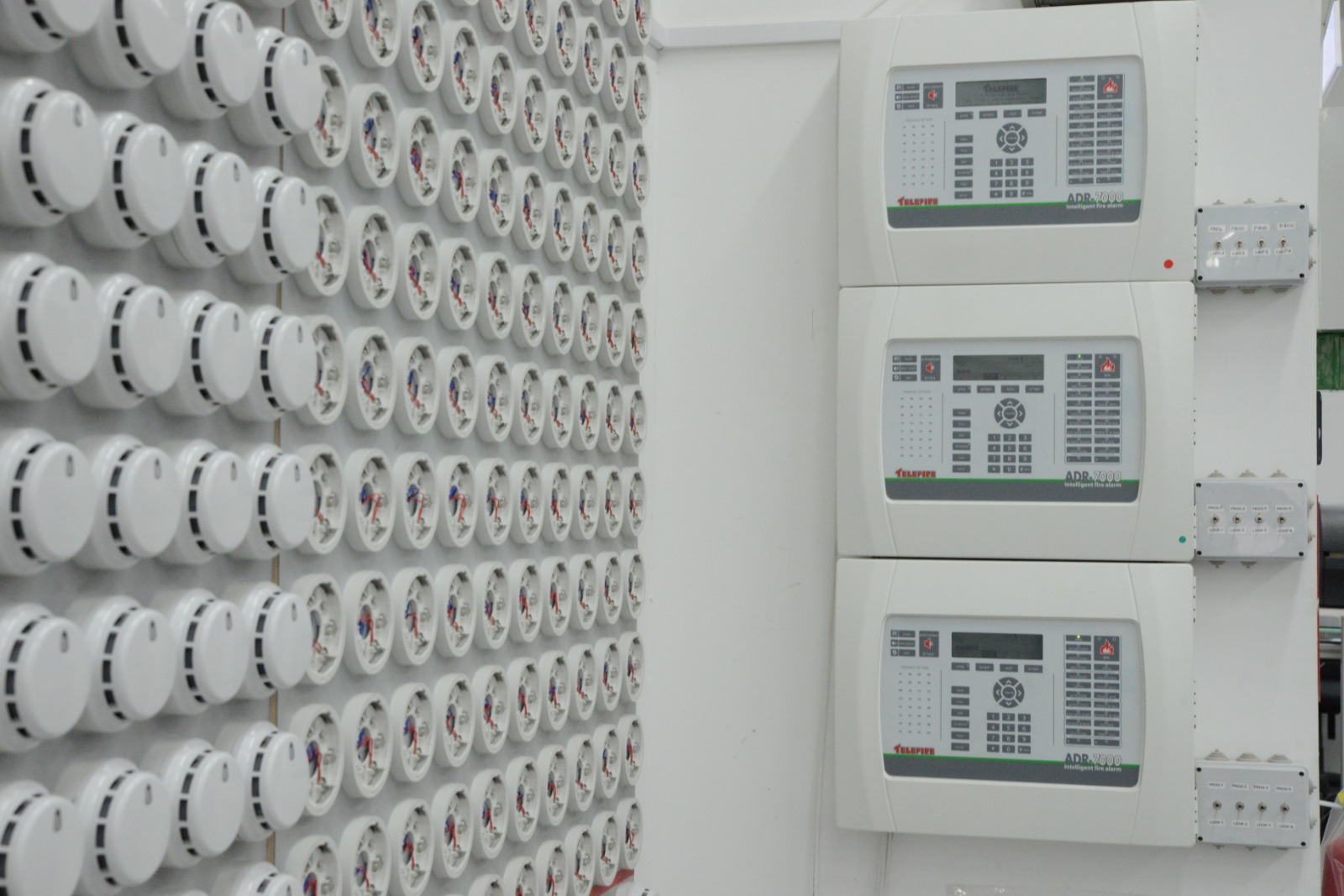 Rows of white ceiling smoke detectors on a wall beside three addressable fire alarm panels.
