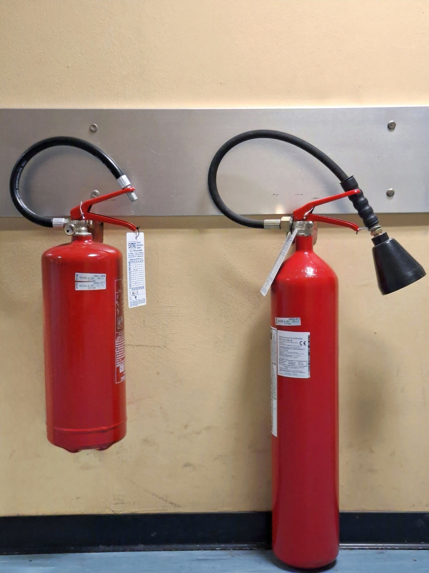 Two red fire extinguishers hanging on a steel wall bracket, one with a discharge horn for carbon dioxide.
